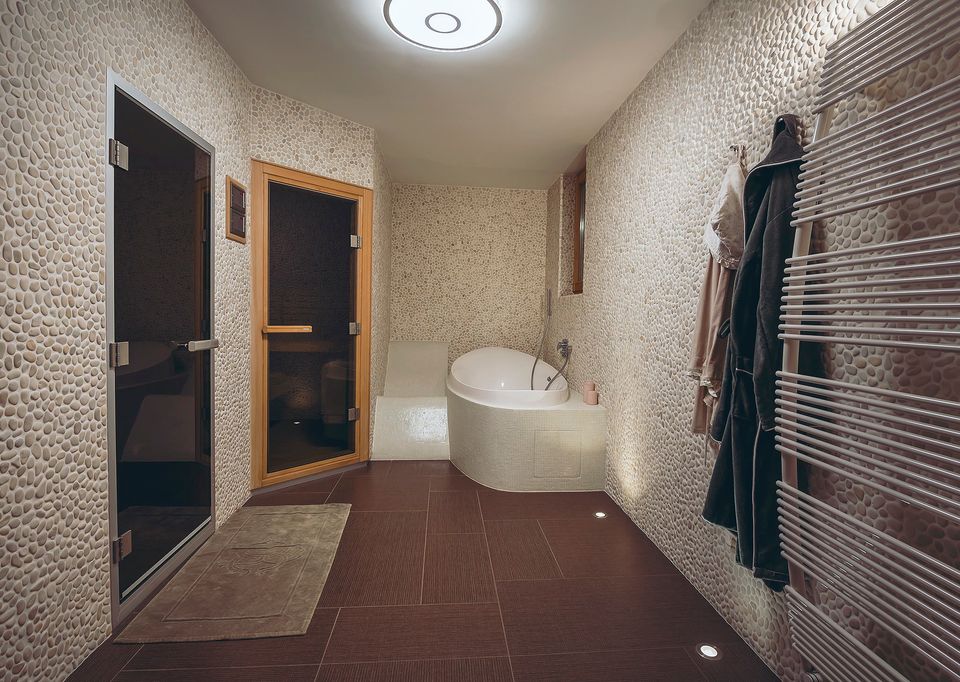 The spa area with a freestanding soaking tub, robes, a heated towel rail, a dark tiled floor, and sauna doors beyond.