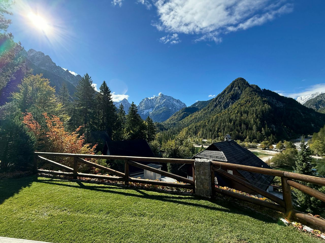The lawn and timber railing in full sun, looking across to the snow-dusted peaks of the Julian Alps under a clear blue sky.
